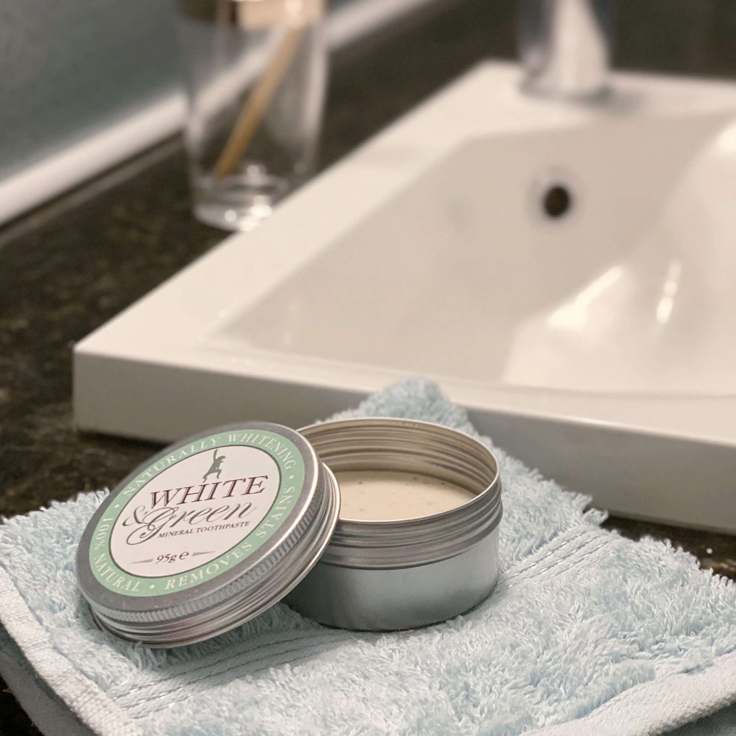 Open tin of White & Green mineral toothpaste on a folded towel beside a bathroom sink, with a toothbrush in the background