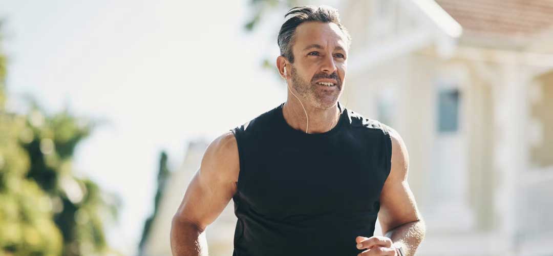Middle-aged man jogging outdoors in the sun wearing a sleeveless top and earphones