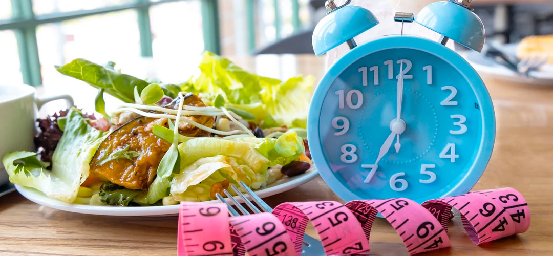 Plate of salad beside a blue alarm clock and pink tape measure, symbolising intermittent fasting