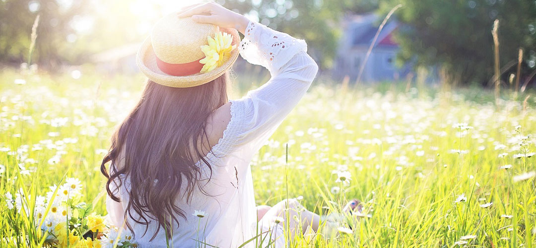 Woman in white dress and straw hat sitting in a sunny wildflower meadow on a summer’s day