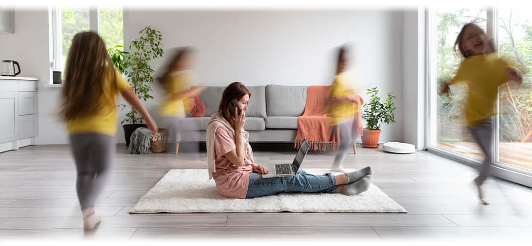 A mum sat on the floor taking a phone call whilst children are running around