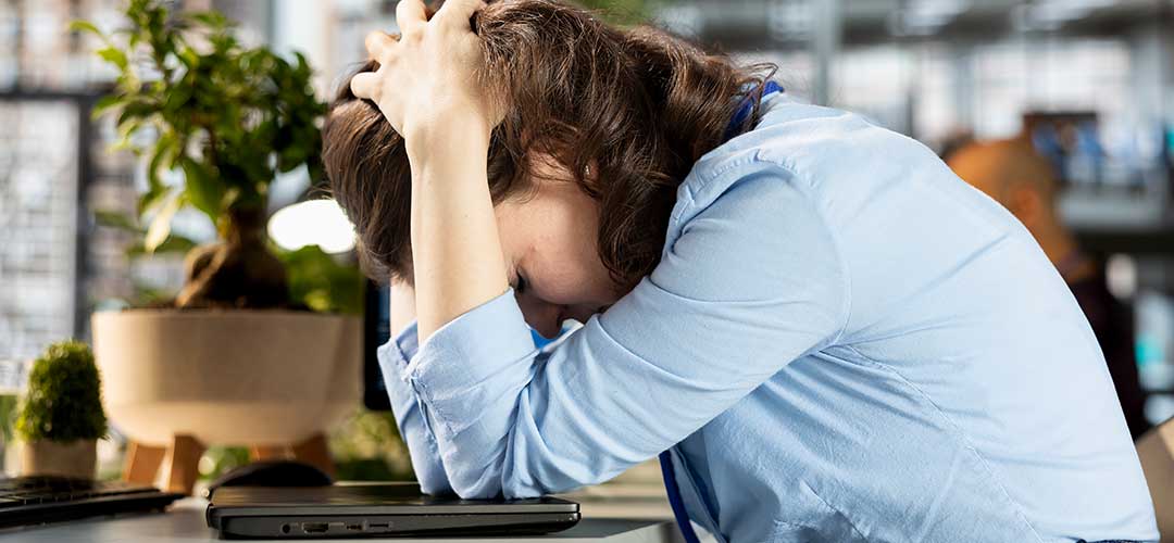Tired woman with head in hands leaning over a laptop at her desk in an office setting.