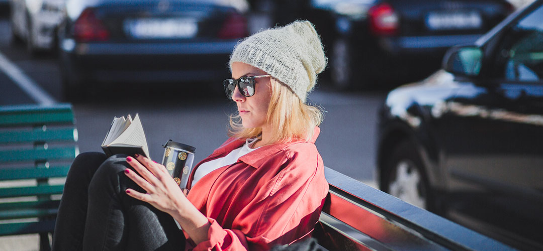 Woman wearing a beanie and sunglasses, sitting on a bench outdoors, reading a book and holding a reusable coffee cup