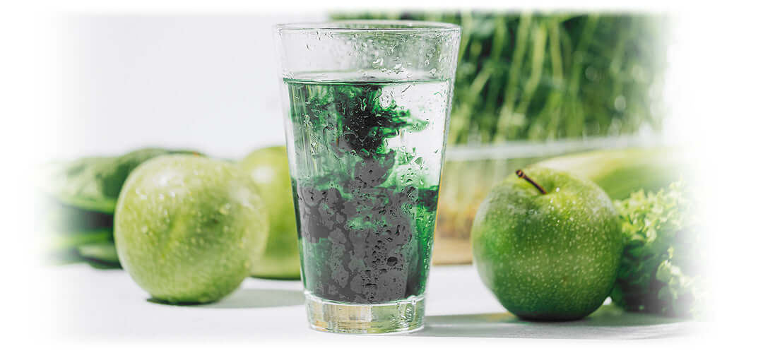 A glass of water with green liquid dispersing, the glass is surrounded by green apples