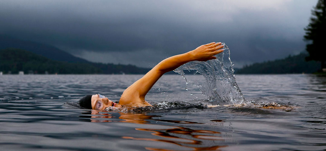 Person swimming in a cold, open lake under cloudy skies, highlighting the UK’s growing wild swimming trend and the benefits of cold water therapy