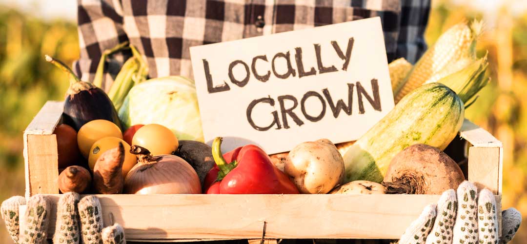 Person holding a wooden crate of UK-grown vegetables with a sign reading "Locally Grown"