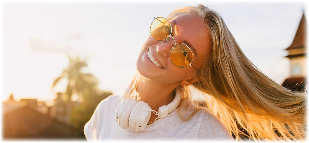 Smiling woman in sunlight wearing sunglasses and headphones, symbolising how supplements and lifestyle habits support a radiant, youthful glow