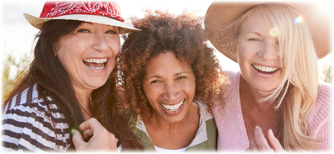 Three joyful women in casual clothing and sun hats laughing together outdoors on a sunny day.