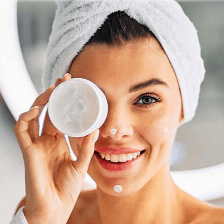 Smiling woman with a towel on her head holding a cream jar to her eye, with small dots of cream on her face
