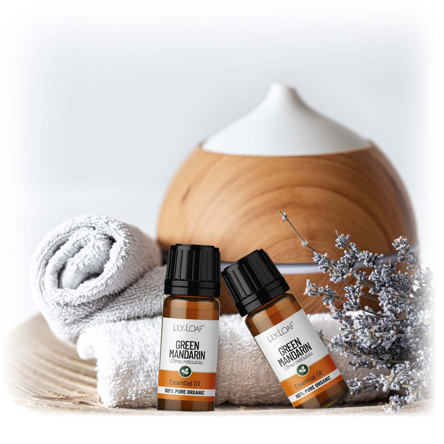 Two bottles of Green Mandarin essential oil with a diffuser, towel, and decorative tree on a light background.