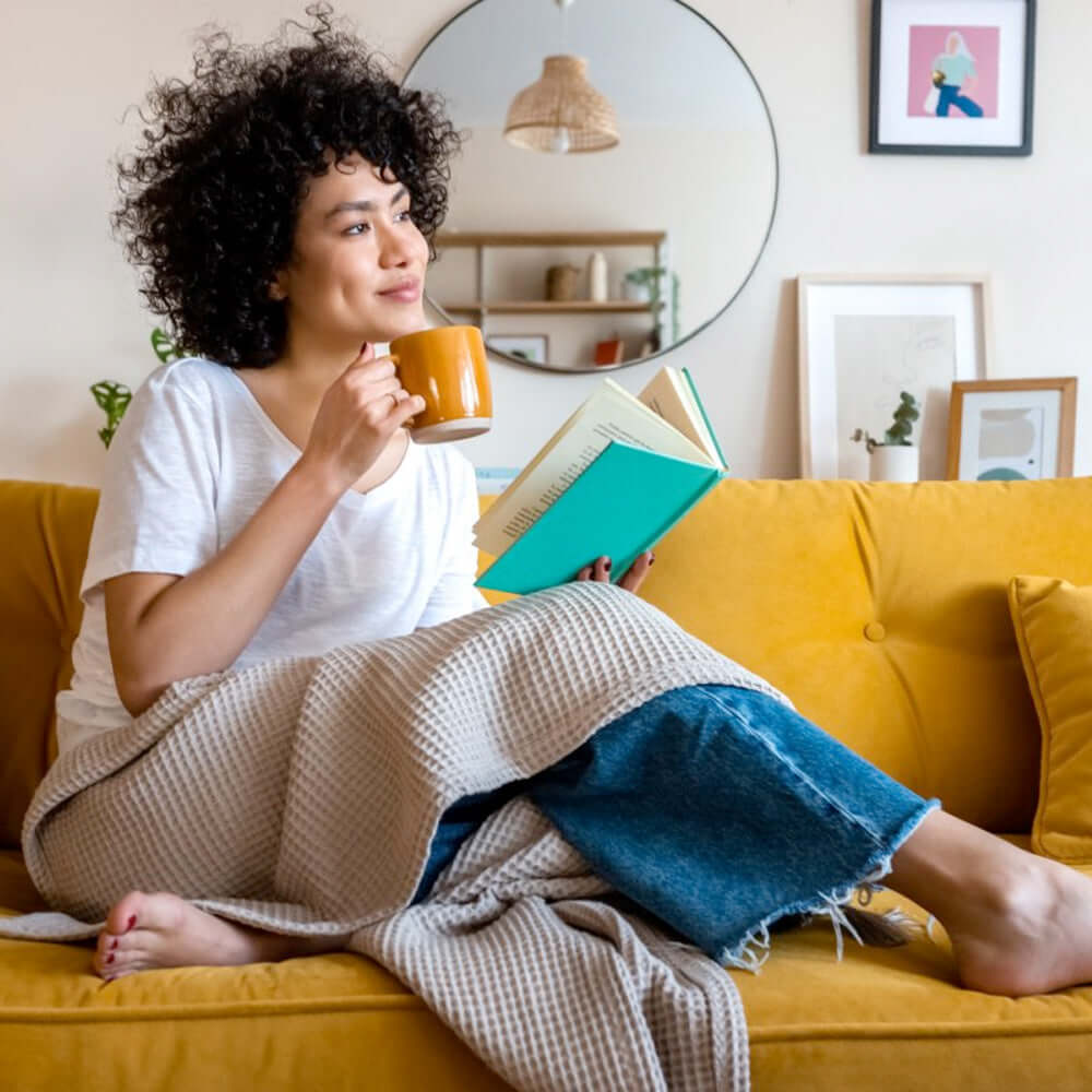 A woman relaxing on a yellow sofa drinking tea and reading a book