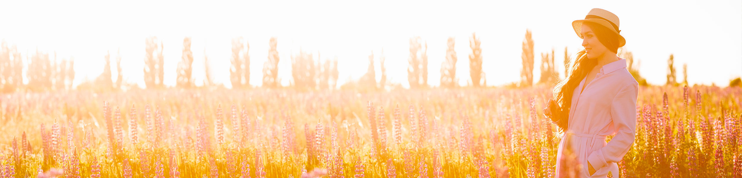 A woman with long hair, wearing a straw hat, in a field of foxgloves at sunset