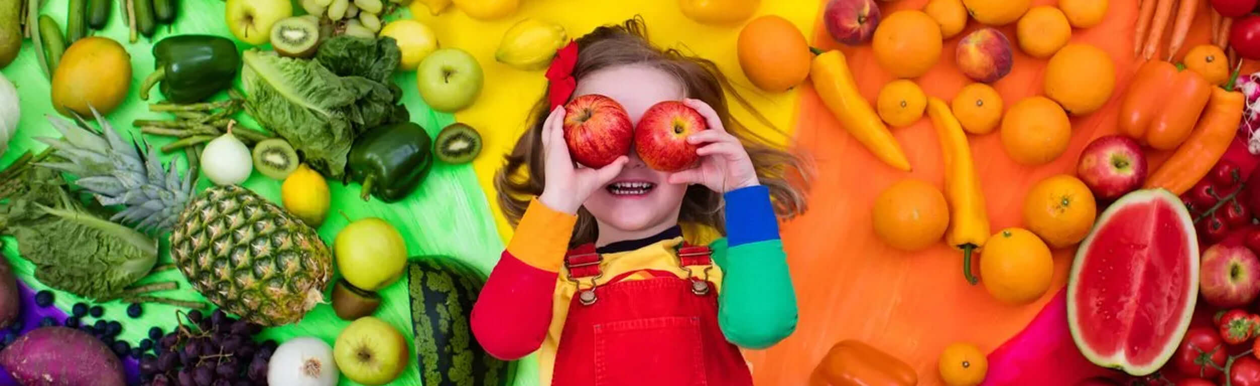 A small girl holding apples against her eyes with a variety of fruits and vegetables on the wall behind her.