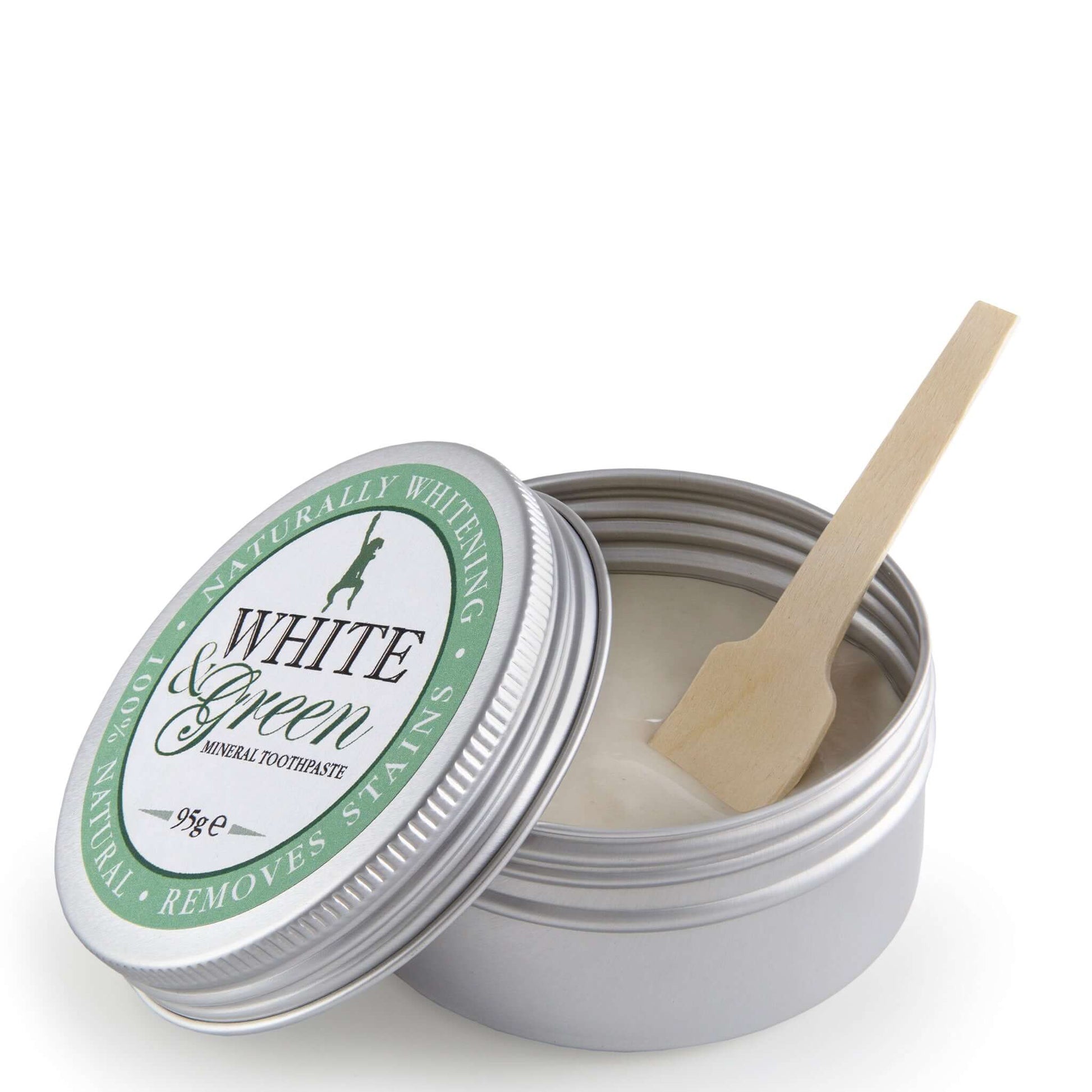 Open tin of White & Green mineral toothpaste with silver lid, green label, and wooden spatula resting inside the creamy paste
