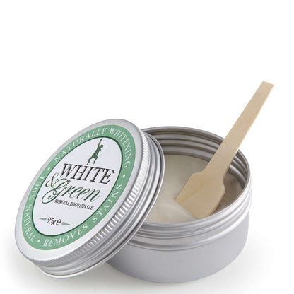 Open tin of White & Green mineral toothpaste with silver lid, green label, and wooden spatula resting inside the creamy paste
