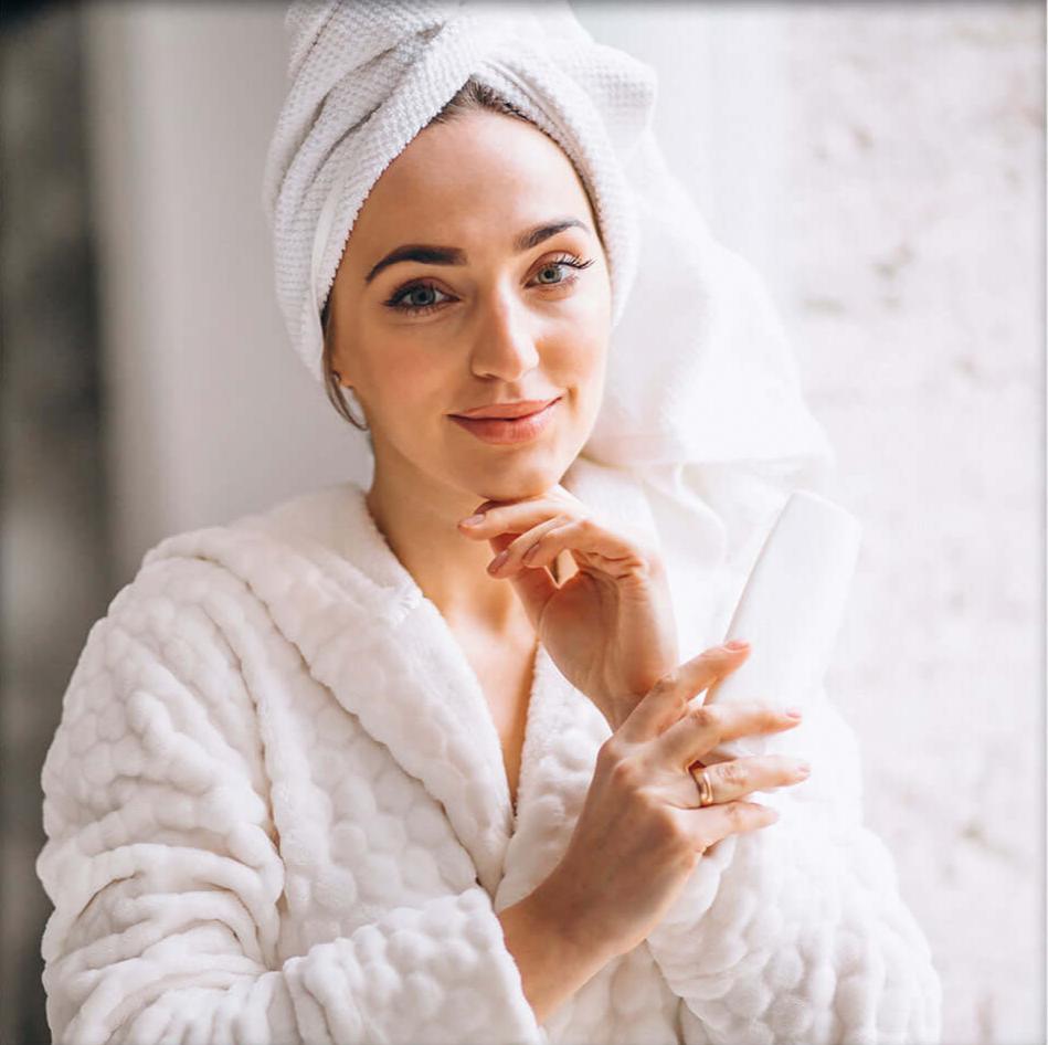 A woman with smooth, glowing skin wearing a towel robe is looking at the camera with a serene expression. 