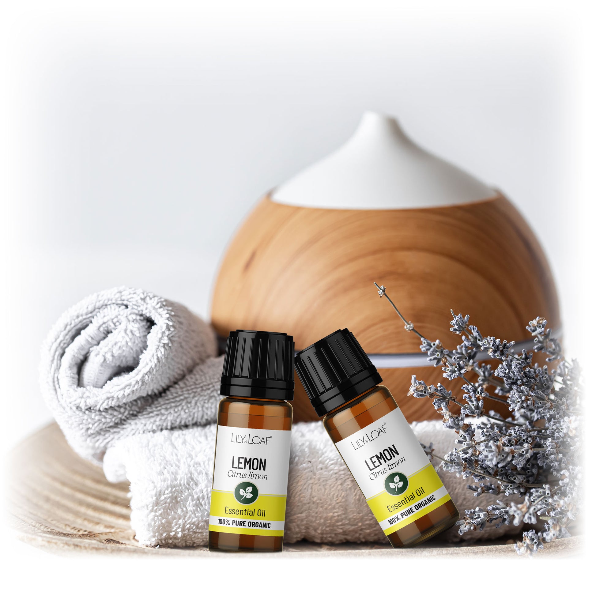 Two bottles of Lily & Loaf Organic Lemon Essential Oil bottle resting on fluffy white towels with a diffuser in the background